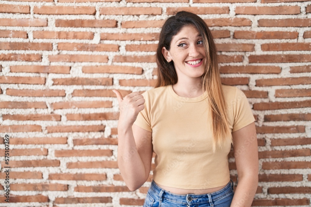 Wall mural Young brunette woman standing over bricks wall smiling with happy face looking and pointing to the side with thumb up.