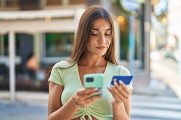 Young beautiful hispanic woman using smartphone and credit card with serious expression at street