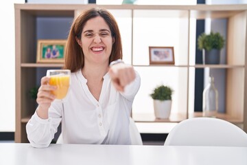 Brunette woman drinking glass of orange juice pointing to you and the camera with fingers, smiling positive and cheerful