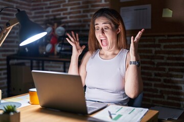 Brunette woman working at the office at night celebrating crazy and amazed for success with arms...