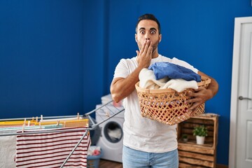 Young hispanic man with beard holding laundry basket covering mouth with hand, shocked and afraid...