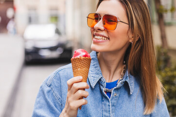 Close up happy young woman with delicious pink ice cream in waffle cone outdoors. Girl wear orange...