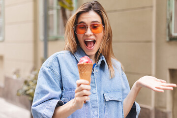 Happy young woman with delicious pink ice cream in waffle cone outdoors. Girl wear orange...