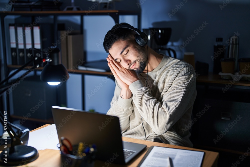 Wall mural Young handsome man working using computer laptop at night sleeping tired dreaming and posing with hands together while smiling with closed eyes.