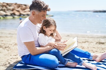 Father and daughter reading book sitting on sand at beach