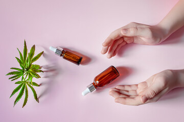Women's hands holding cosmetic bottles on a pink background. Fresh hemp leaves to make elexir for cosmetics