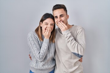 Young hispanic couple standing over white background laughing and embarrassed giggle covering mouth with hands, gossip and scandal concept