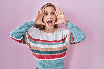 Young blonde woman standing over pink background smiling cheerful playing peek a boo with hands showing face. surprised and exited