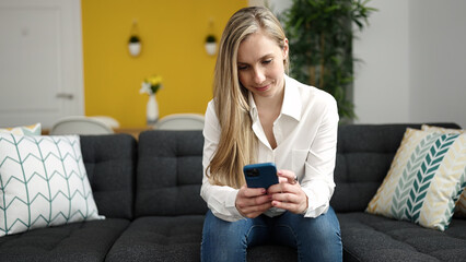 Young blonde woman using smartphone sitting on sofa at home