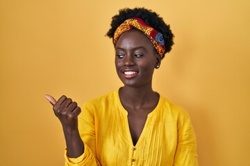African young woman wearing african turban smiling with happy face looking and pointing to the side with thumb up.