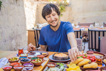 Man eating turkish breakfast. Turkish breakfast table. Pastries, vegetables, greens, olives, cheeses, fried eggs, spices, jams, honey, tea in copper pot and tulip glasses, wide composition - obrazy, fototapety, plakaty