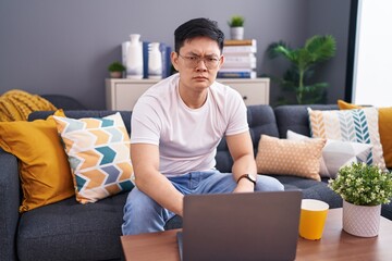 Young asian man using laptop at home sitting on the sofa skeptic and nervous, frowning upset because of problem. negative person.
