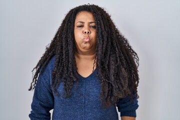 Plus size hispanic woman standing over white background puffing cheeks with funny face. mouth...