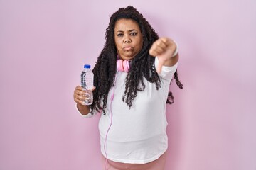 Plus size hispanic woman wearing sportswear and headphones looking unhappy and angry showing rejection and negative with thumbs down gesture. bad expression.