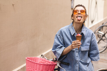 Happy laughing blonde girl with high ponytail in denim shirt and short , top outfit wear orange sunglasses and hold pink ice cream on a summer day next to a bright bike fixed. Girl show tongue. - Powered by Adobe