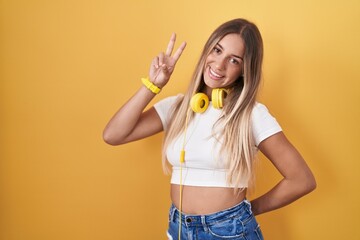 Young blonde woman standing over yellow background wearing headphones smiling looking to the camera showing fingers doing victory sign. number two.