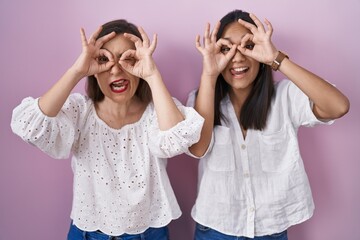 Hispanic mother and daughter together doing ok gesture like binoculars sticking tongue out, eyes looking through fingers. crazy expression.