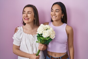 Hispanic mother and daughter holding bouquet of white flowers looking away to side with smile on face, natural expression. laughing confident.