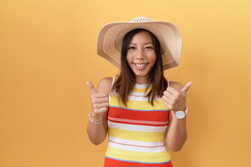 Middle age chinese woman wearing summer hat over yellow background success sign doing positive gesture with hand, thumbs up smiling and happy. cheerful expression and winner gesture.