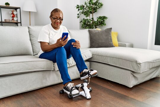 Senior african american woman using smartphone and doing sport using bike pedals machine at home