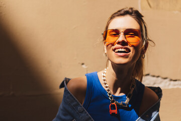 Attractive woman with high ponytail standing near beige wall on the street of old city. Girl joy of sun light, wear orange sunglasses look at sun. Shadow and light of sunny summer, spring or fall day. - obrazy, fototapety, plakaty