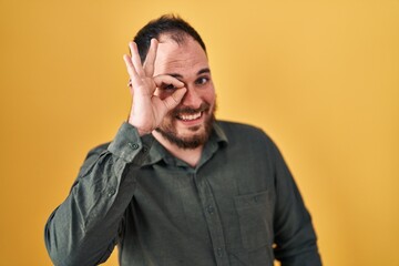 Fototapeta premium Plus size hispanic man with beard standing over yellow background doing ok gesture with hand smiling, eye looking through fingers with happy face.