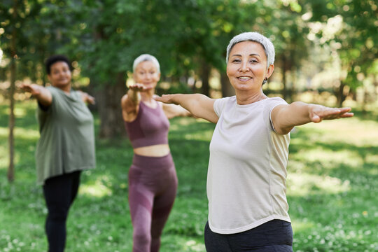 Waist up portrait of smiling senior woman enjoying sports workout outdoors and looking at camera while doing stretching exercise, copy space