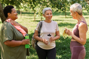Waist up portrait of three senior women chatting after outdoor workout in park and holding water bottles