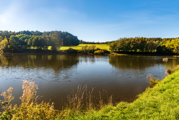 Early autumn rural landscape with pond, trees, meadows, and blue sky