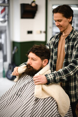 Man covered with haircut cape sits in hairdressers chair and barber covers his shoulders with towel.