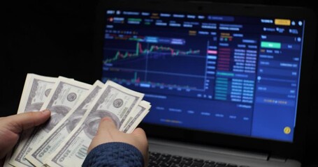 A man counts one hundred dollar bills against the background of a cryptocurrency chart on a laptop screen. A man earns money by trading bitcoin on a cryptocurrency exchange.