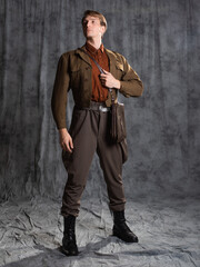 A young guy in military-style clothes, a brown flight jacket and breeches with suspenders. Posing in the studio on a gray background - 575142770