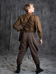 A young guy in military-style clothes, a brown flight jacket and breeches with suspenders. Posing in the studio on a gray background, back view