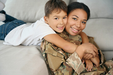 Mixed race boy tenderly hugs his mother, woman in military uniform