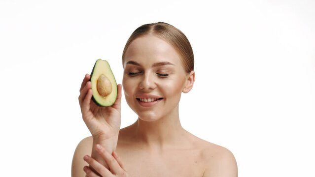 Close up shot of tender blue eyed woman looks directly at camera holds halves of avocado near face chooses healthy nutrition has perfect smooth skin isolated over beige background. beauty concept