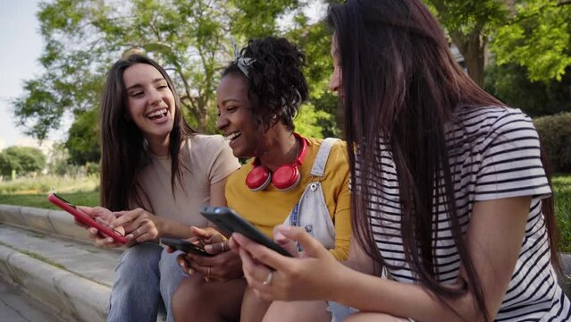 Happy young women using cell phones outdoors. Group smiling college friends checking social networks on smartphone. Classmates having fun watching dating apps on mobile. Young girls flirting online. 