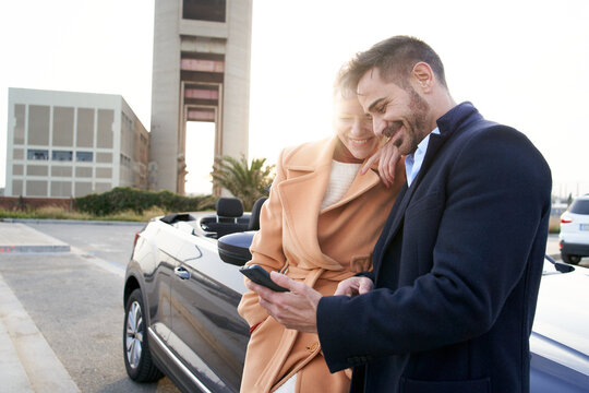 Cheerful Middle-aged Couple Looking At Cell Phone Leaning On Convertible Car. Smiling And Happy Couple Having Fun. Good Moments Of A Couple Relationship. Portrait Of Wife And Husband Outdoors.