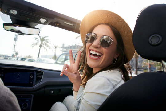 Portrait of pretty young hipster with big smile in convertible car. Beautiful woman with sunglasses and hat making peace sign. People renting car for vacation on a sunny day. Funny moments outdoors.