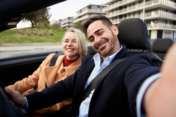 Happy middle-aged couple takes selfie in the car. Wife and husband pose and look smiling at camera....