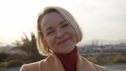 Happy mature blonde woman at sunset looking for camera and touching her hair. Adult enjoying the fresh air whose bob is moved by the wind. Middle aged people posing outside the city.