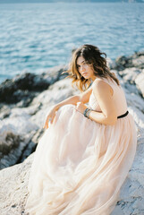 Fototapeta na wymiar Bride in a beige dress with tousled hair sits on the rocks by the sea