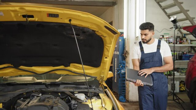 Bearded man car technician mechanic repairing car problem of engine, during system checking detail, using laptop computer for maintenance and fixing in car garage. Repairing car problem of engine.