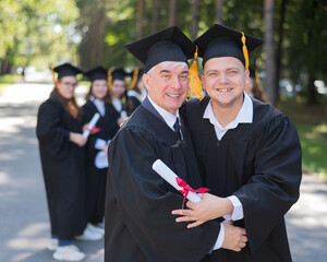 A group of graduates in robes outdoors. An elderly man and a young guy congratulate each other on receiving a diploma.