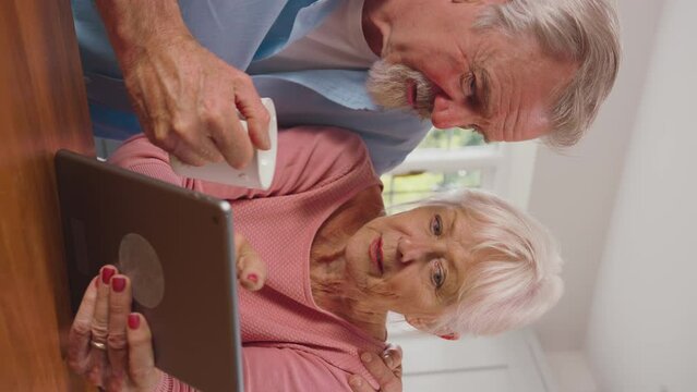 Vertical video of retired senior couple with digital tablet at home in kitchen shopping online or booking holiday - shot in slow motion