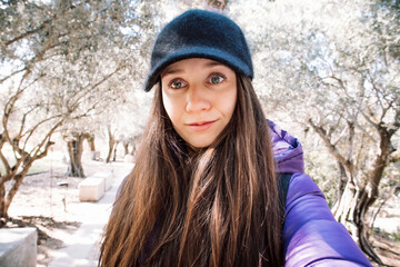 A beautiful young happy woman walking in spring autumn park botanical garden among olive trees plantation. Portrait of a white European girl wearing a black cap smiling looking straight in a camera.