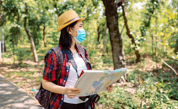 Asian woman tourism traveling in a beautiful green park wearing a protective mask with camera and backpack and hold the map, Asian women looking right side during travel in the forest, Outdoor new nor