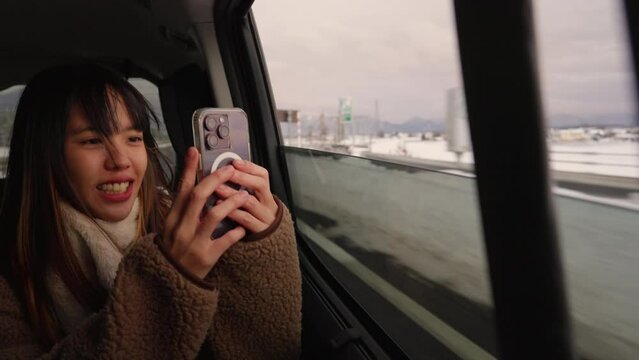 Asian woman traveler using mobile phone taking picture of beautiful view with snow out of the window during travel on car at sunset. Attractive girl enjoy travel road trip in Japan on winter vacation.