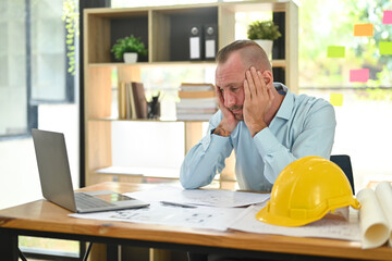 Stressed caucasian engineer man working with blueprints, examining construction plans at his...