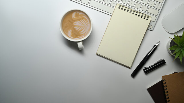 Top view of spiral notepad, pen, cup of coffee and keyboard on white office desk. Copy space for text