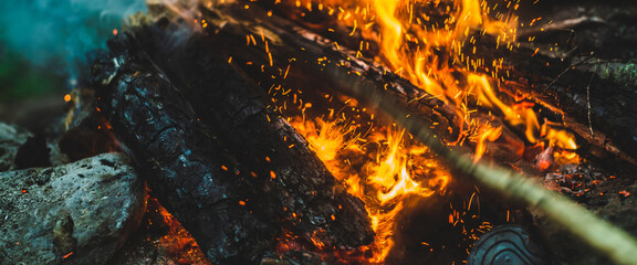 Vivid smoldered firewoods burned in fire close-up. Atmospheric background with orange flame of campfire and blue smoke. Warm full frame image of bonfire with glowing embers in air. Bright sparks bokeh